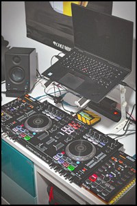 A DJ setup featuring a laptop elevated on one of the latest DJ Laptop Stands, a speaker, and a professional DJ controller with multiple buttons, knobs, and jog wheels on a desk.