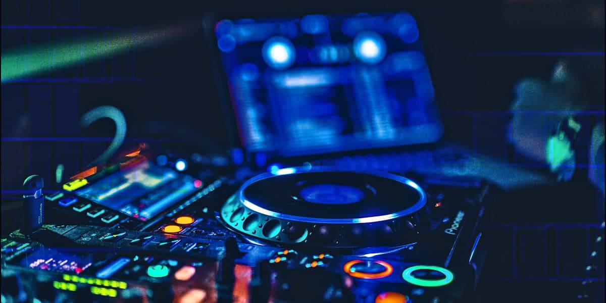 Close-up of a DJ mixing console and turntable with colorful lights, hinting at Mobile DJ Basics, with a blurred computer screen in the background.