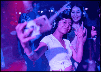 Two young women wearing headphones take a selfie at a silent disco party, dancing to their favorite school dance songs with colorful lights in the background.