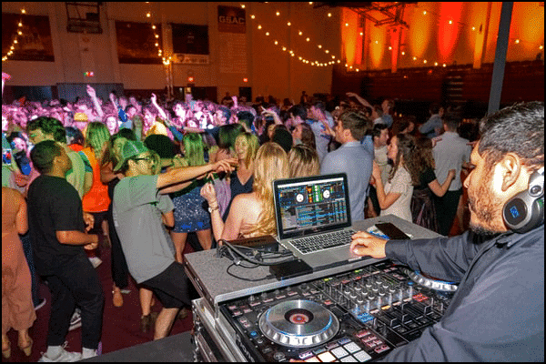 A DJ plays School Dance Songs from a laptop as a large group of people dance and socialize on a crowded, brightly lit dance floor.