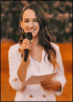 A woman in a white blazer holds a microphone and paper, smiling while speaking outdoors—perfectly embodying tips on How To MC a Wedding with confidence and charm.