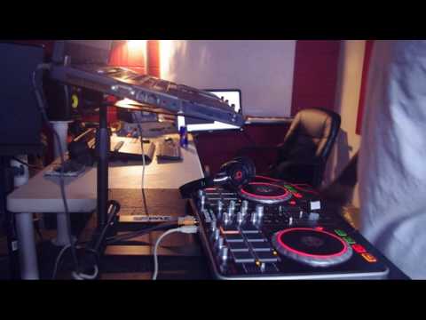 A DJ controller and headphones sit on a desk in a music studio, with DJ laptop stands, a chair, computer monitor, and keyboard visible in the background.