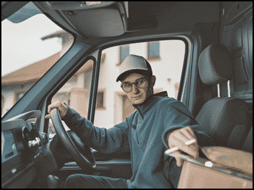 A person wearing glasses and a cap sits in the driver’s seat of a vehicle, holding a package with one hand and the steering wheel with the other—perhaps delivering equipment in one of the top vehicles for DJs.