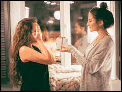 Two women indoors; one covers her face in surprise while the other presents a small gift box with a ribbon, perfect for those searching for unique Gift Ideas for DJs.