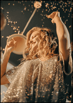 A woman in a sparkling dress smiles and raises her arms as confetti falls around her at a party, celebrating to her favorite party songs.