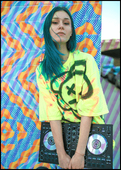 Person with blue hair wearing a neon yellow shirt with a bear graphic holds a DJ controller, standing in front of a colorful, geometric patterned background—showcasing What Should a Mobile DJ wear to stand out at any event.