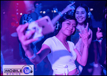 Two young women at a silent disco event take a selfie, wearing wireless headphones. Neon lights and other partygoers are visible in the background, while a "Mobile DJ Basics" logo hints at must-have School Dance Songs.
