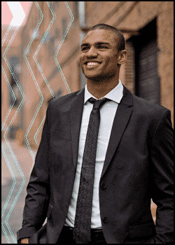 A man in a dark suit and tie stands outside in an urban setting, smiling, with geometric lines overlaid on the left side—a great example of what should a mobile DJ wear for a polished, professional look.