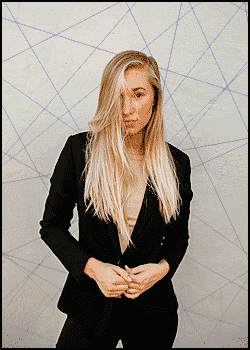 A person with long blonde hair wearing a black blazer stands in front of a white wall decorated with intersecting blue lines, showcasing a stylish look perfect for those wondering what should a mobile DJ wear.
