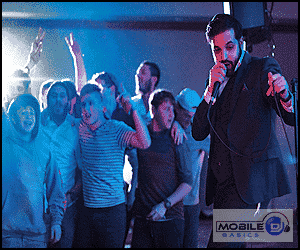 A DJ in a suit holds a microphone while a group cheers and poses behind him at an indoor event under blue lighting, ready to answer any questions to ask clients about their music preferences.
