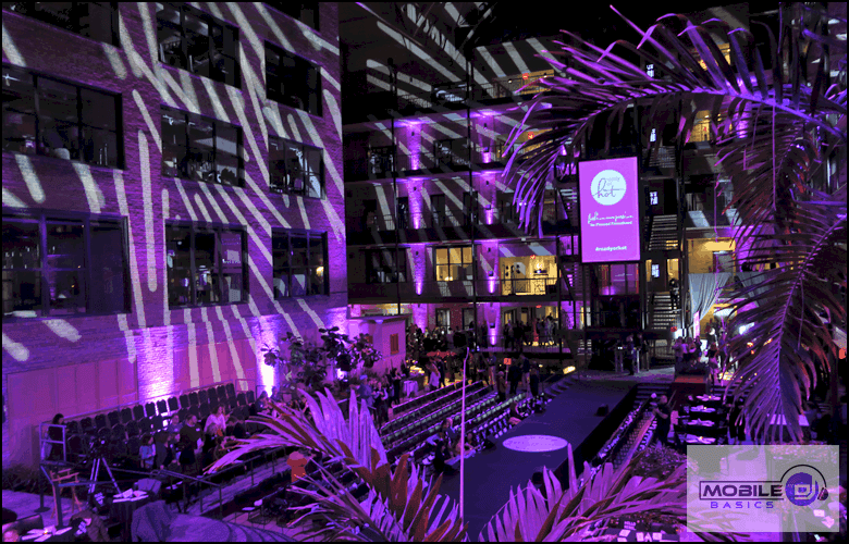 An outdoor event space with rows of seating and a runway is illuminated by purple and white DJ lighting, with projected logos on the building and decorative plants in the foreground.