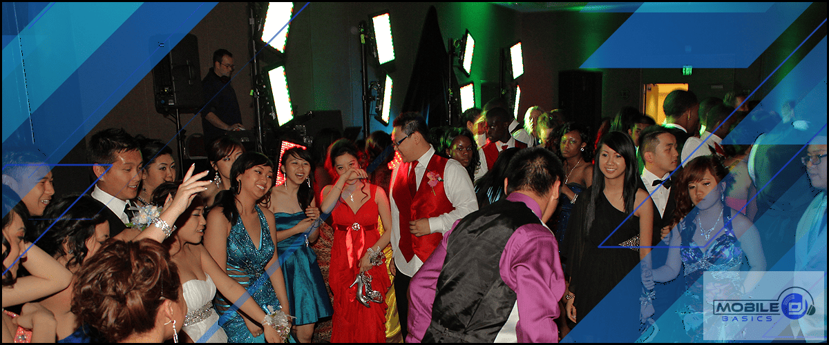 A large group of people in formal attire dance and socialize at an indoor event with colorful lighting and a DJ setup by Ryan Conlon.