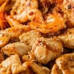 Close-up of a plate with grilled shrimp, chicken pieces, sliced vegetables, and rice, garnished with sesame seeds.