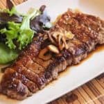 Grilled steak topped with sliced garlic, served with a side of mixed greens on a rectangular white plate.