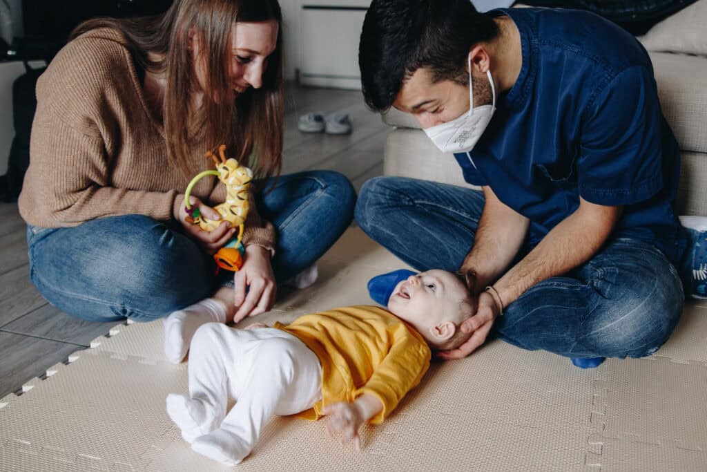 Foto di Lorenzo mentre visita un neonato: il bimbo è sdraiato sul tappetone, Lorenzo gli sorregge la testa e la mamma del bimbo gli sta a fianco.