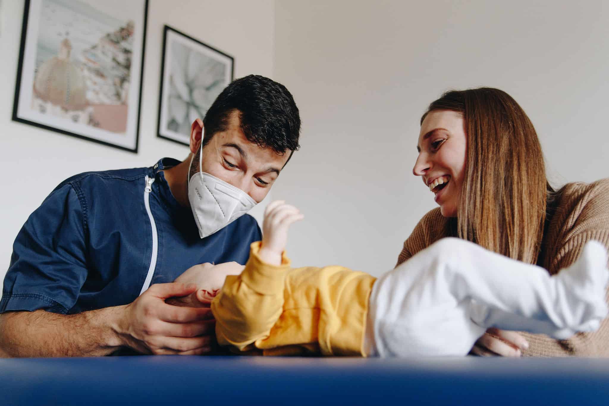 Foto di Lorenzo mentre visita un bimbo sul lettino, con a fianco la mamma che sorride
