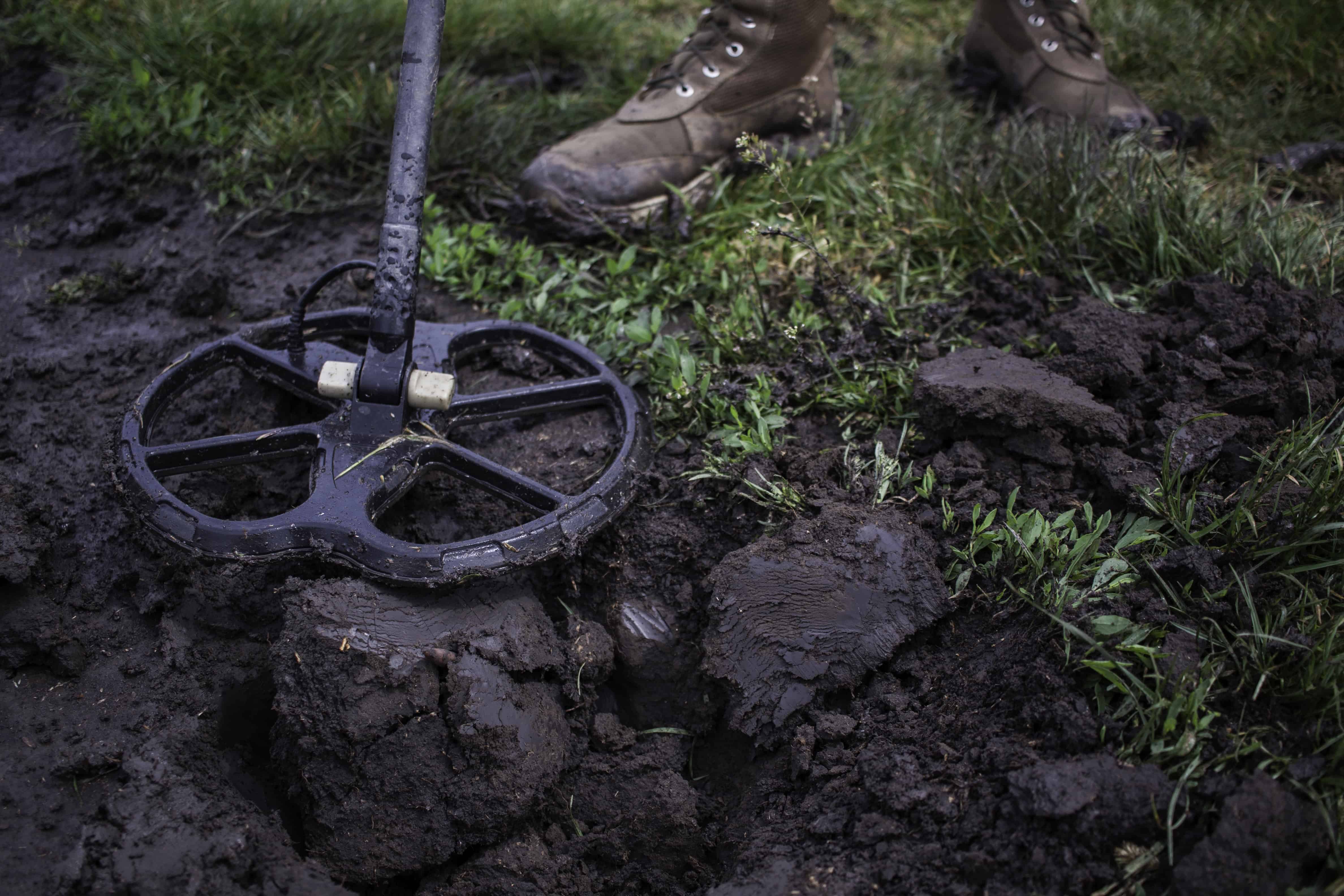 A person searching for detecting gold nuggets uses a metal detector to scan muddy ground, with patches of grass and boots visible in the scene.