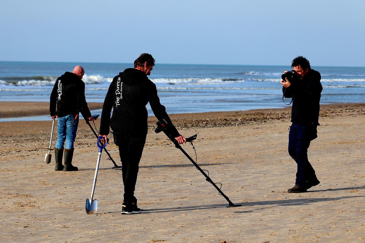 Three people on a sandy beach; two demonstrate how metal detectors work with shovels, while the third person takes a photo. The ocean is visible in the background.