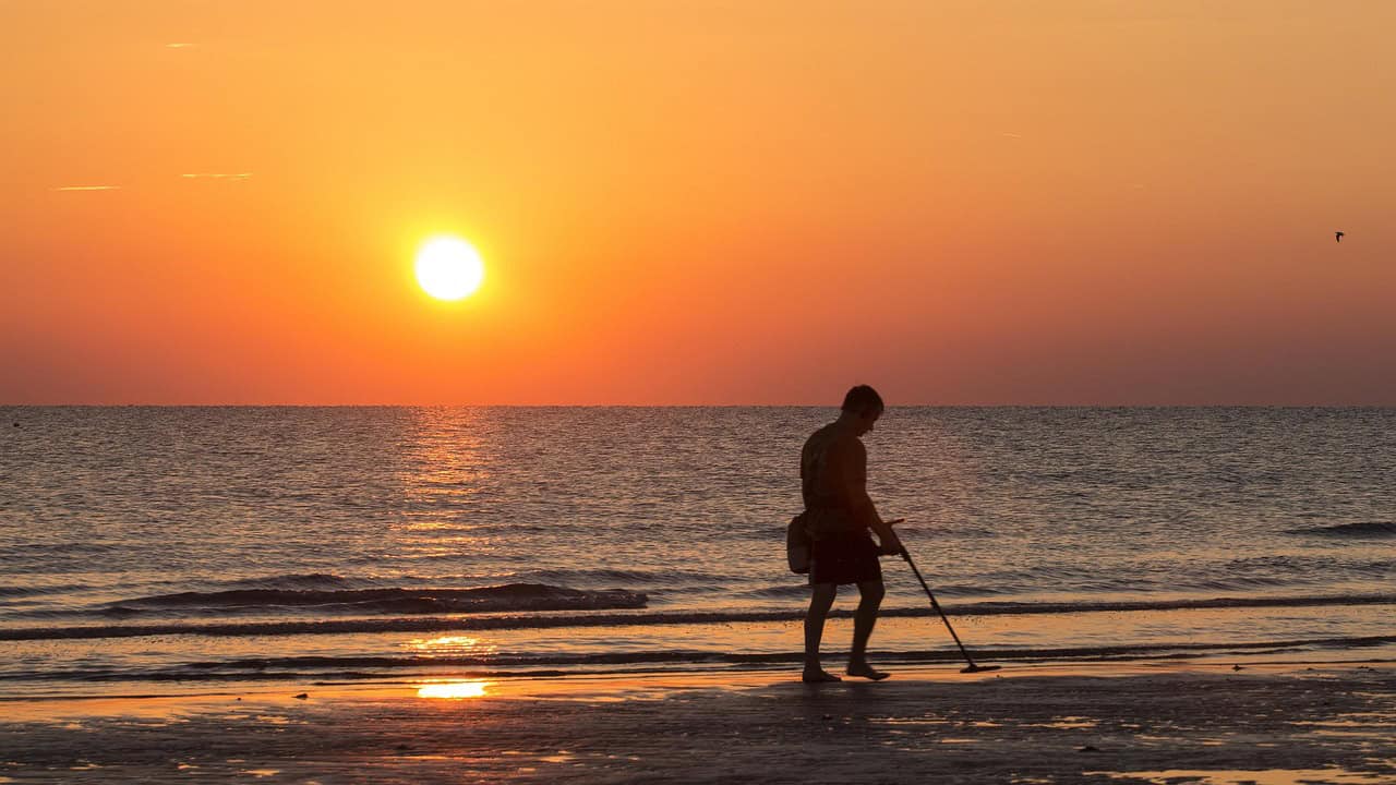 A person follows Metal Detecting Laws and Regulations while using a metal detector on the beach at sunset, with the ocean and sun low in the sky.