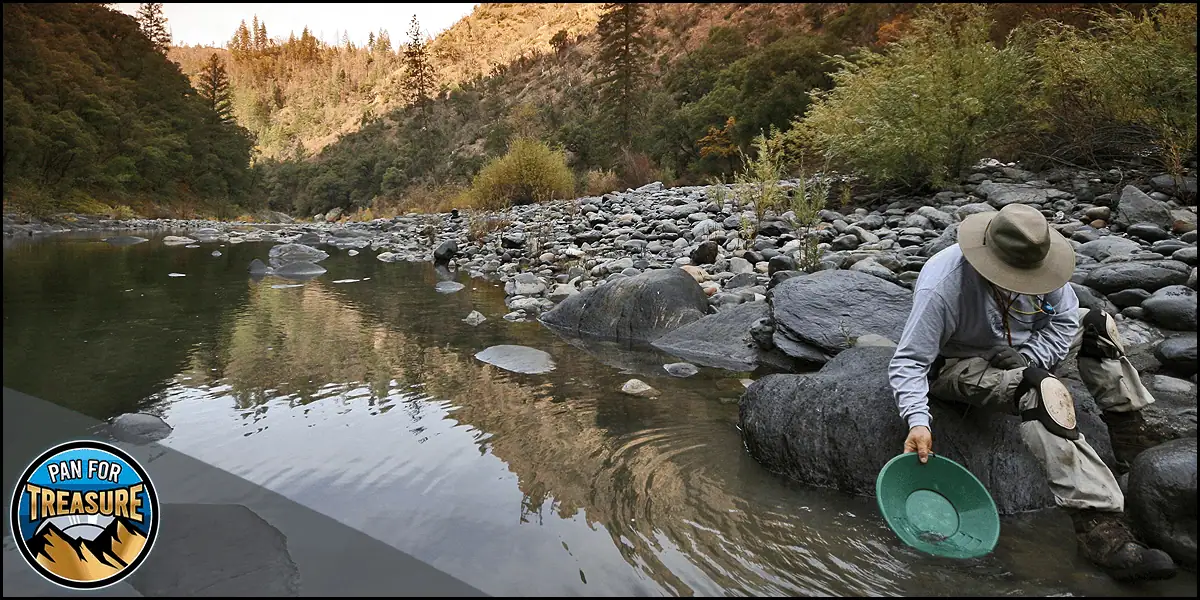 A man in a hat is kneeling on rocks next to a river panning for gold - Gold panning in a riverbed with prospecting tools