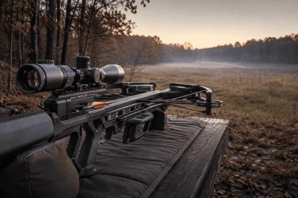 Crossbow fitted with a variable-power scope on a shooting bench beside autumn woodland