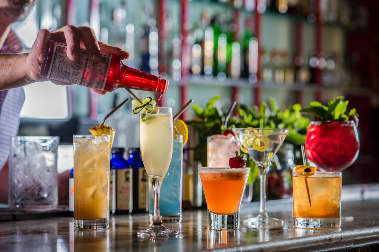 A bartender pouring red syrup into a row of colorful drinks along a bar