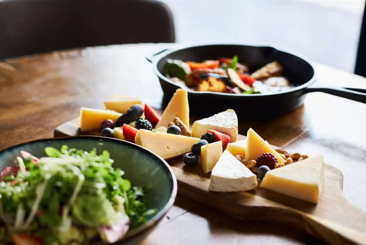 Cold salad, room-temperature cheese board, and hot cast-iron vegetables on wood table