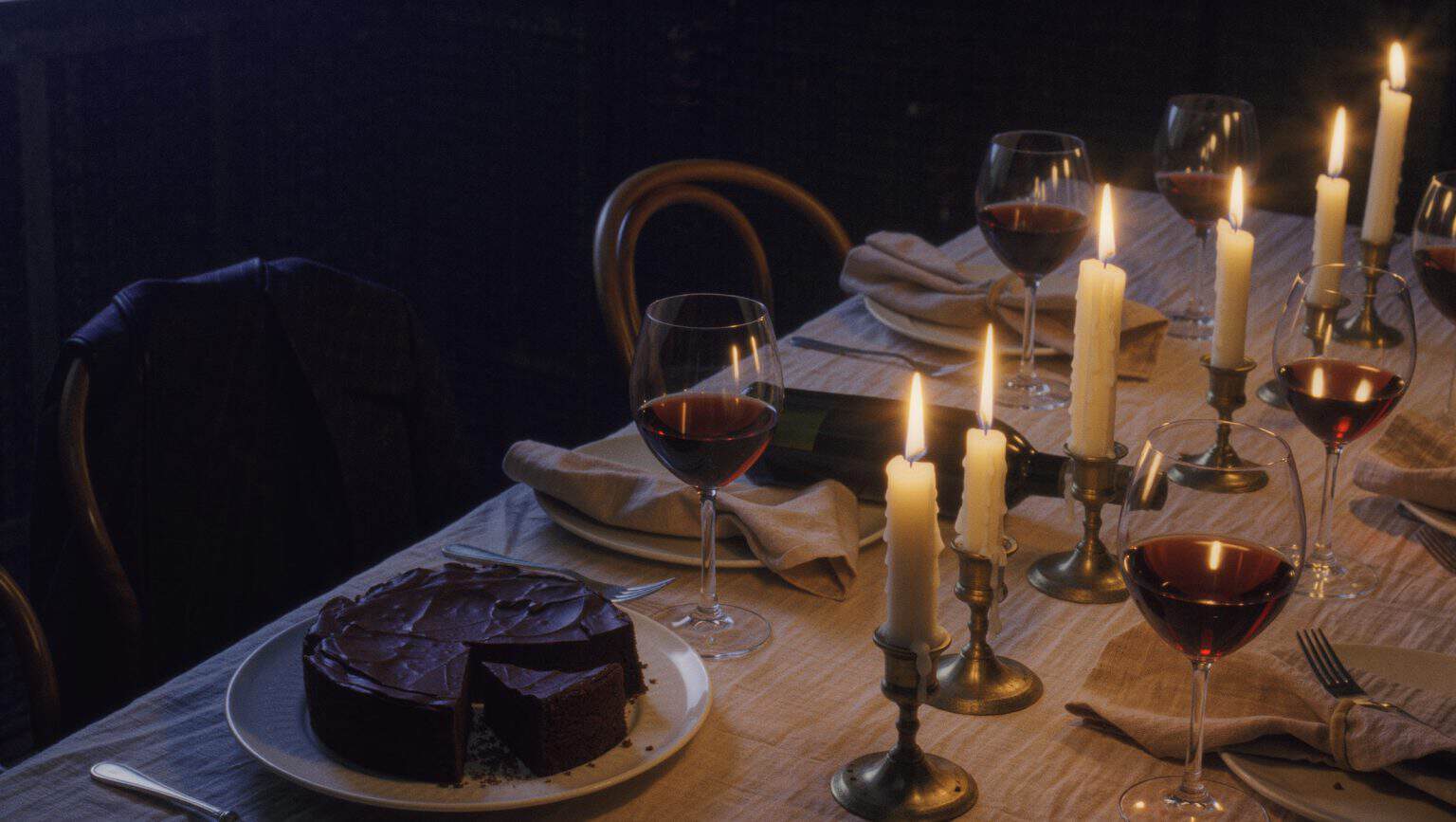 A dinner table mid-evening: candles burned low with running wax, wine glasses at different levels, a chocolate cake with a cracked top and two slices gone, crumpled napkins and chairs pushed back
