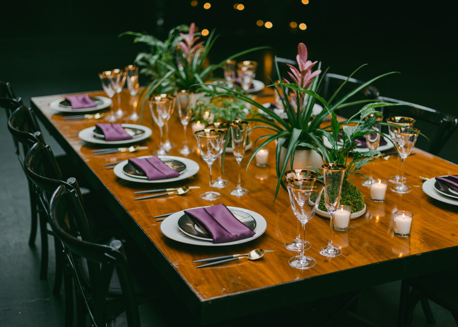 A dark dinner table set with glassware and tall green arrangements