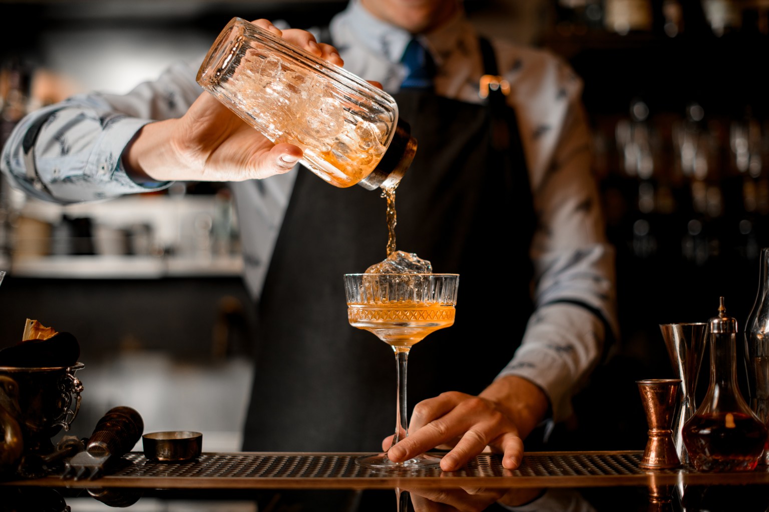 A bartender pouring an amber cocktail into a ribbed glass at a bar