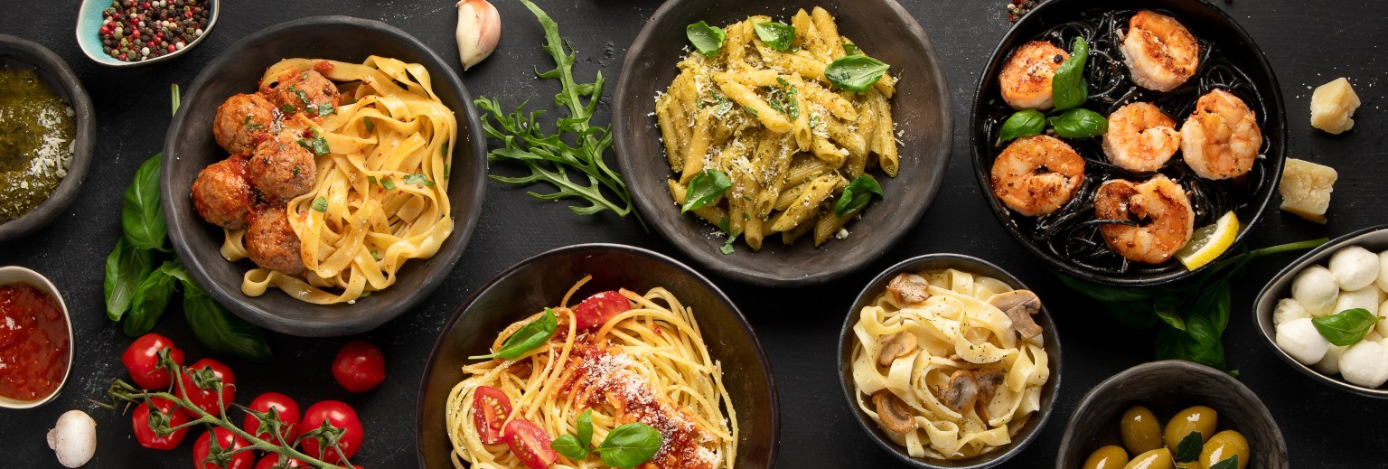 Several bowls of different pastas viewed from above on a dark surface