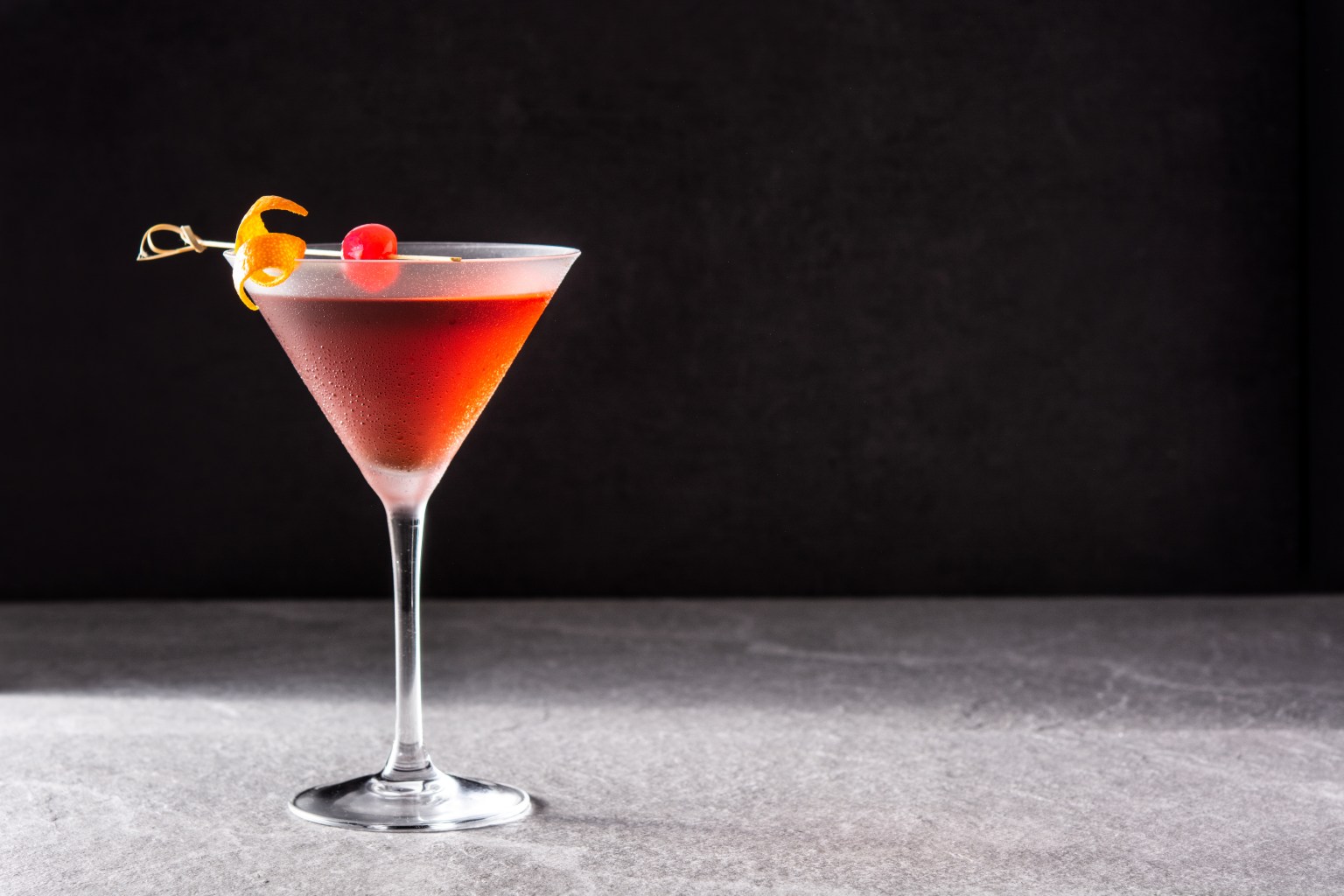 A red martini with a cherry garnish against a plain dark background