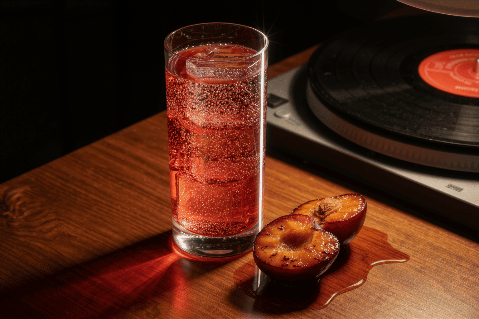 A tall reddish highball full of bubbles on teak beside roasted plum halves and a turntable