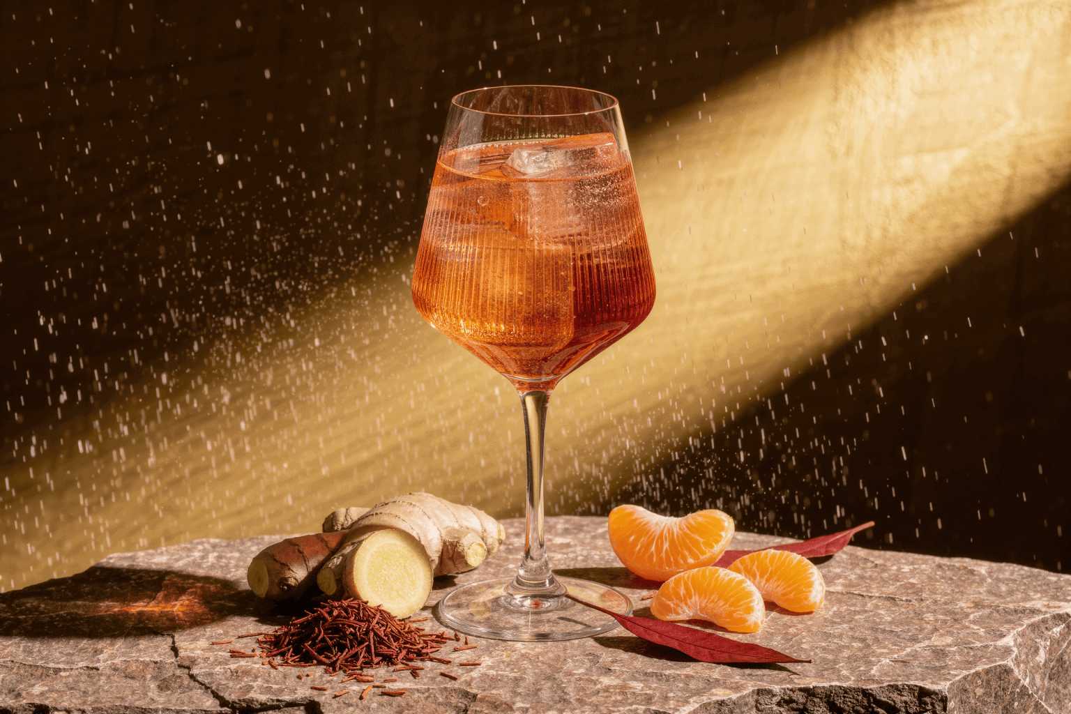An amber spritz backlit on warm sandstone with fresh ginger, mandarin segments, and loose rooibos