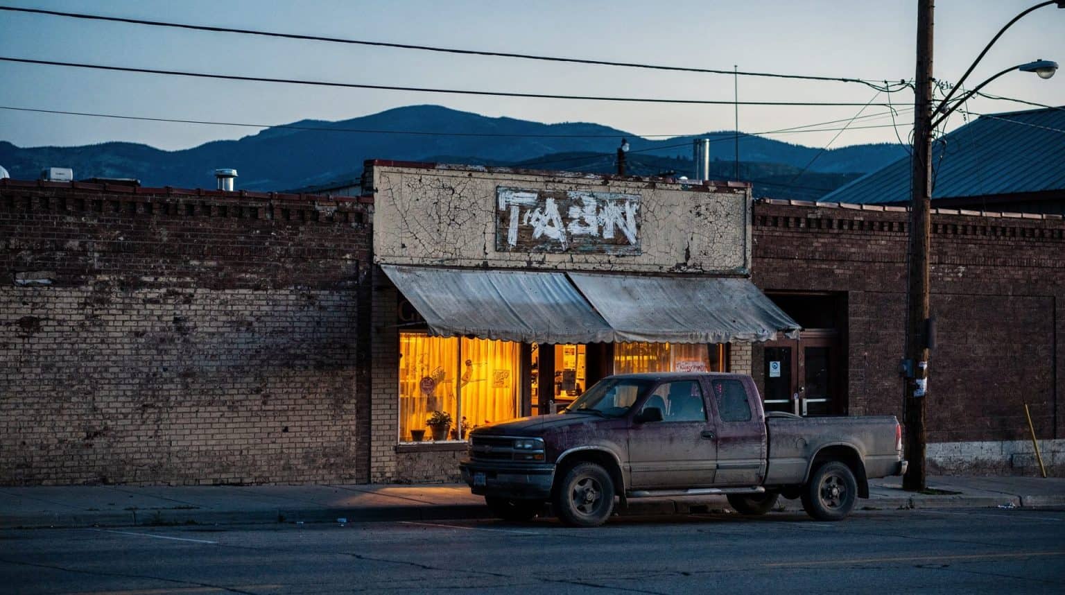 A quiet Montana main street at dusk, low storefronts with a lit restaurant window and blue mountains rising behind the roofline