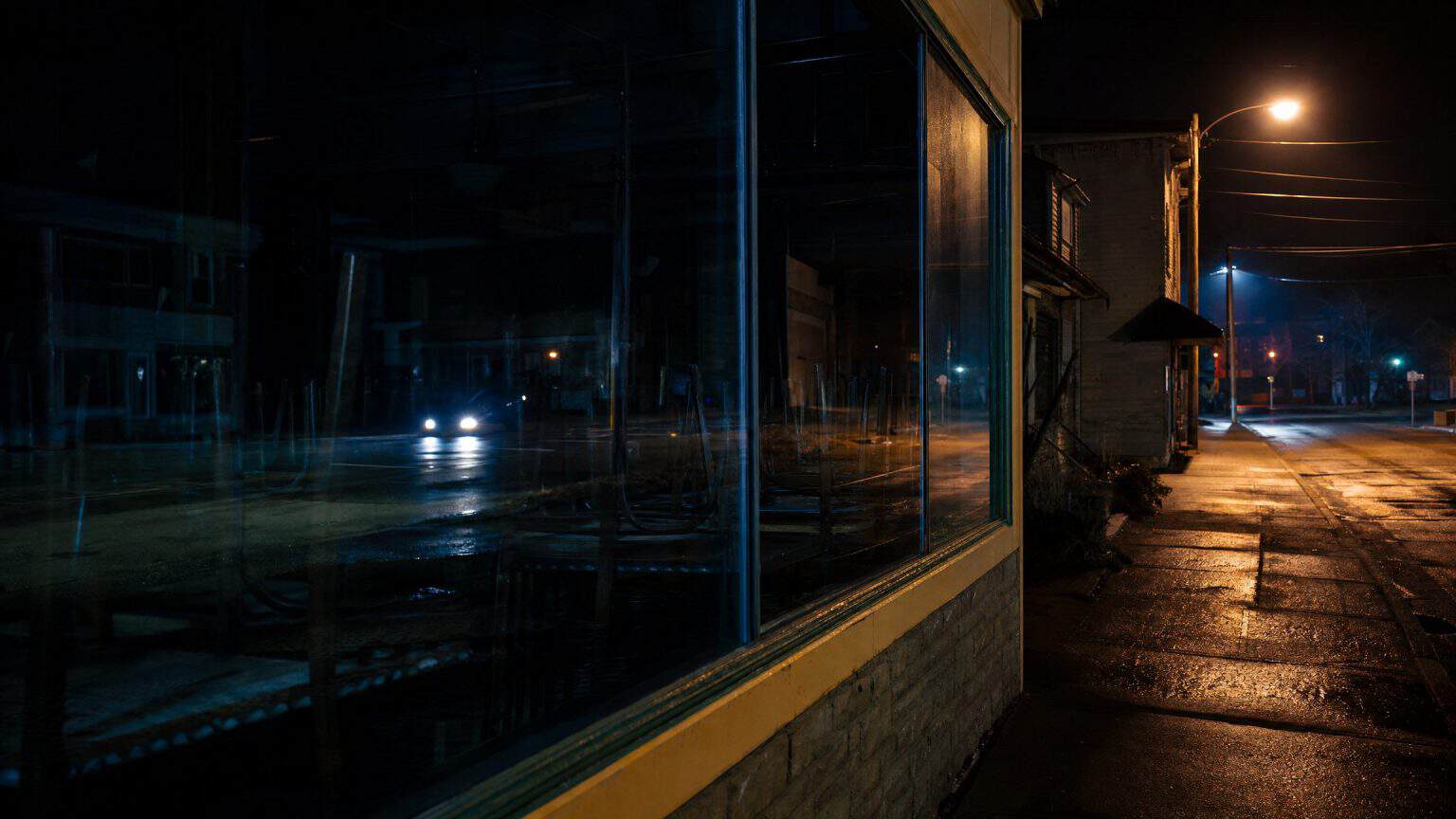 A darkened restaurant window on a quiet small-town street at night, chairs upturned on tables inside, car headlights reflected across the glass.