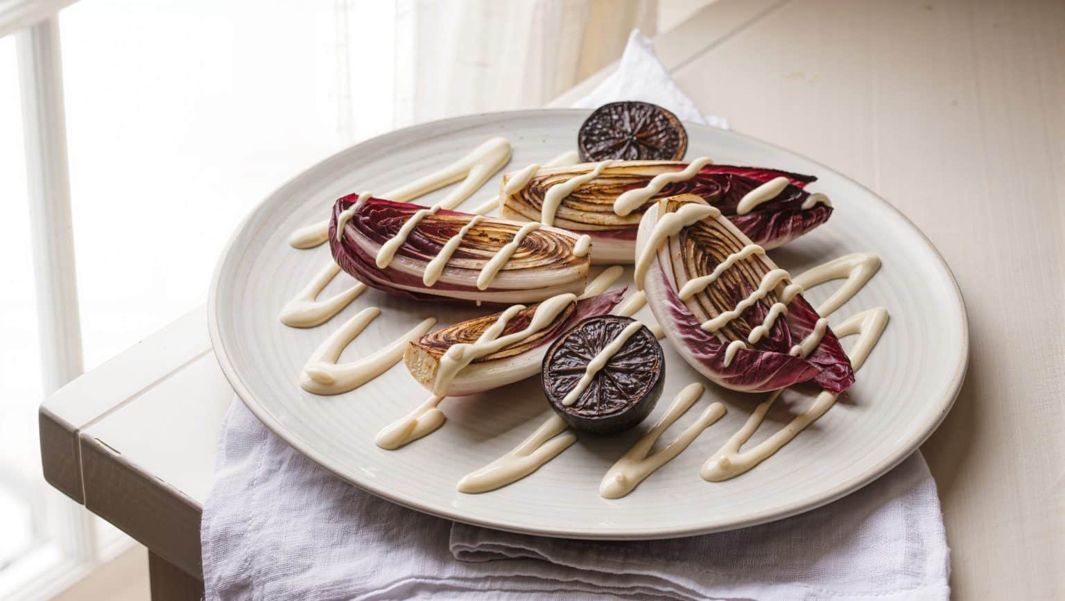 Charred radicchio wedges on a pale platter with streaks of whipped cream and blackened lemon halves, in bright daylight.