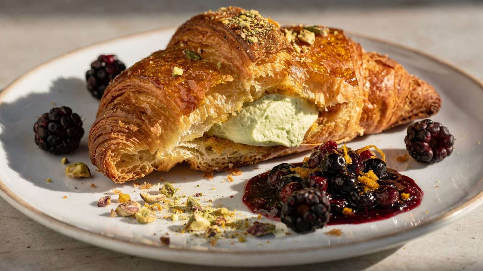 Brûléed croissant French toast showing pistachio filling beside glossy blackberry-orange compote and chopped pistachios.