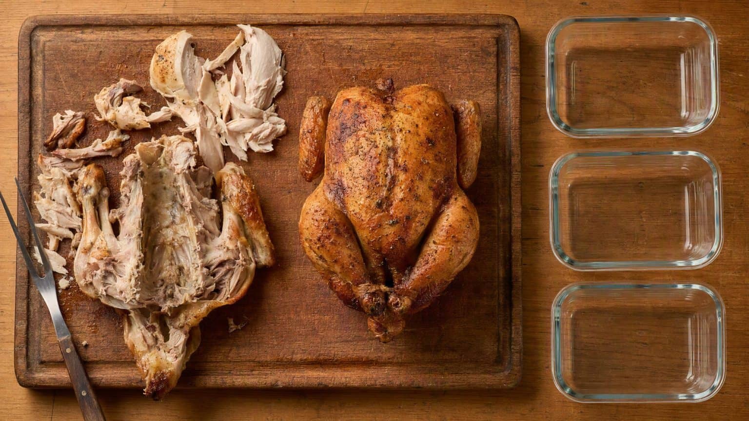 Two whole roast chickens on a wooden board seen from above, one carved down to the frame and one untouched, with three empty glass storage containers lined up beside them.