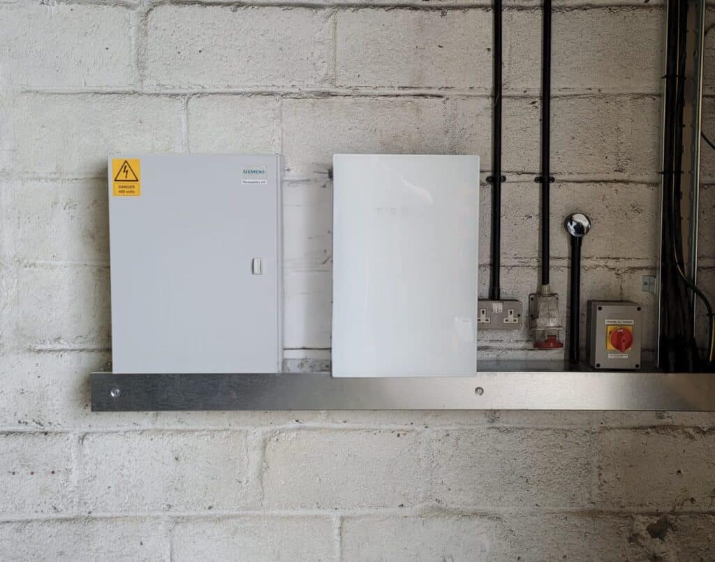 A Siemens electrical panel and a Tesla Powerwall are mounted side by side on a concrete wall with several visible cables and power outlets.