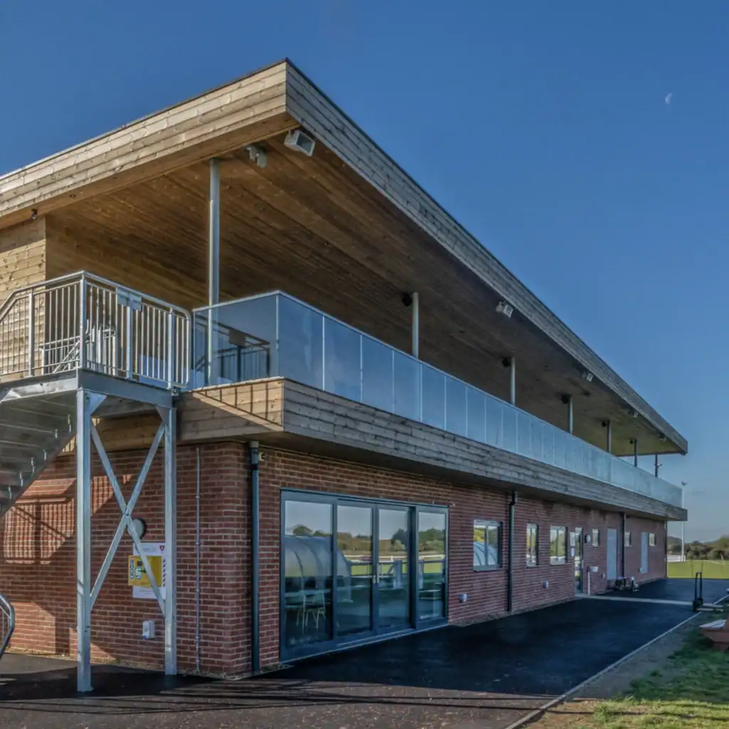 A two-story sports facility with a brick ground floor, wooden upper floor, external staircase, glass balcony, and a shield logo on the wall, under a clear blue sky.
