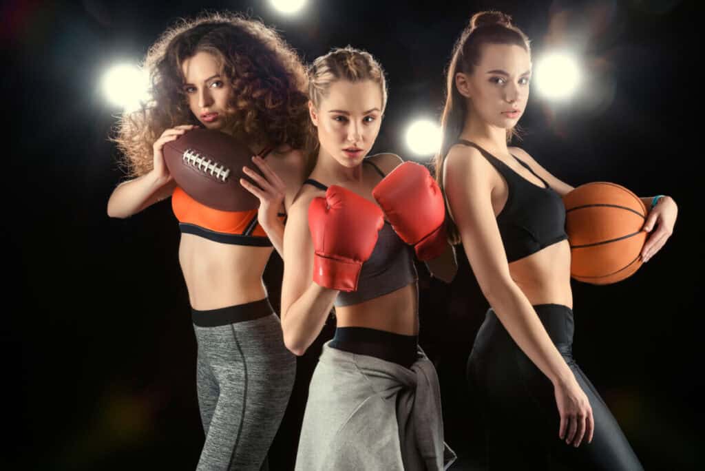 3 sporty women standing next to each other holding a basketball, football and one wearing boxing gloves.