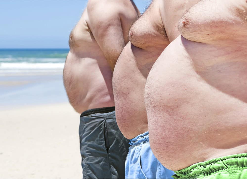 three shirtless men standing on a beach with fatty chests. 