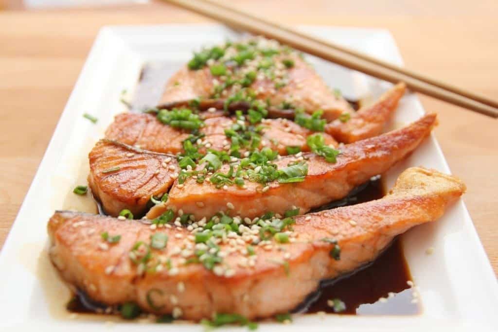 a rectangle white dinner plate with 3 salmon filets laying in a soy sauce with spices on top