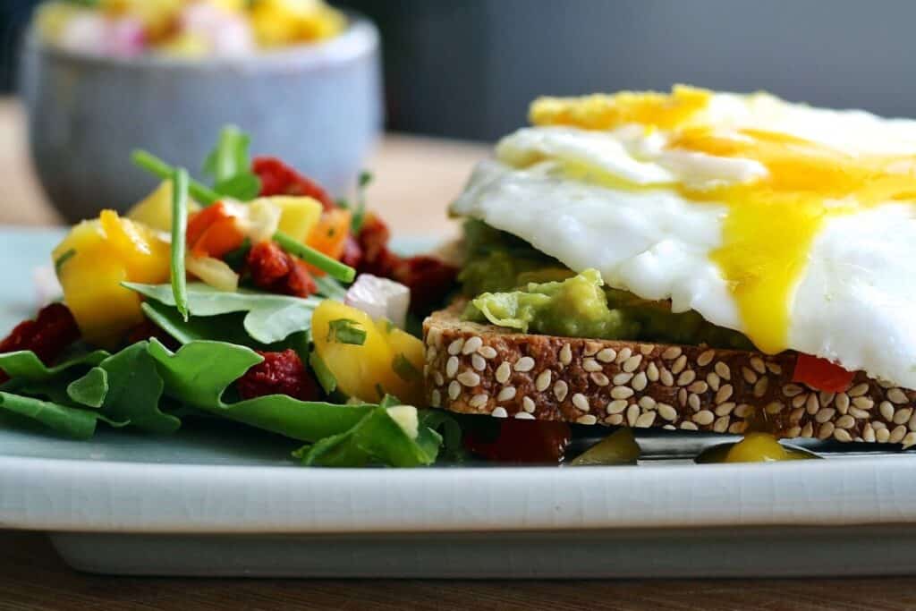 Ezekiel bread with avocado, eggs, and onions making a sandwich. Fruit salad next to the sandwich on a plate.