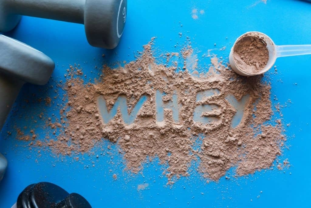 spilled whey protein with the word whey spelled out in the middle, on a blue background with blues dumbbells and a protein scoop on the side.