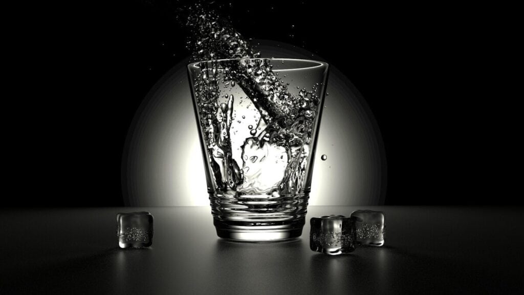 A glass with water being poured in it on a black background sitting on a black table. 3 ice cubes on the table around the glass.