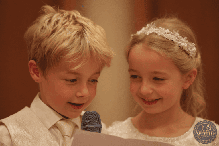 Two young children dressed formally, with a boy holding a microphone and a sheet of paper, stand closely together and appear to be speaking or reading aloud.