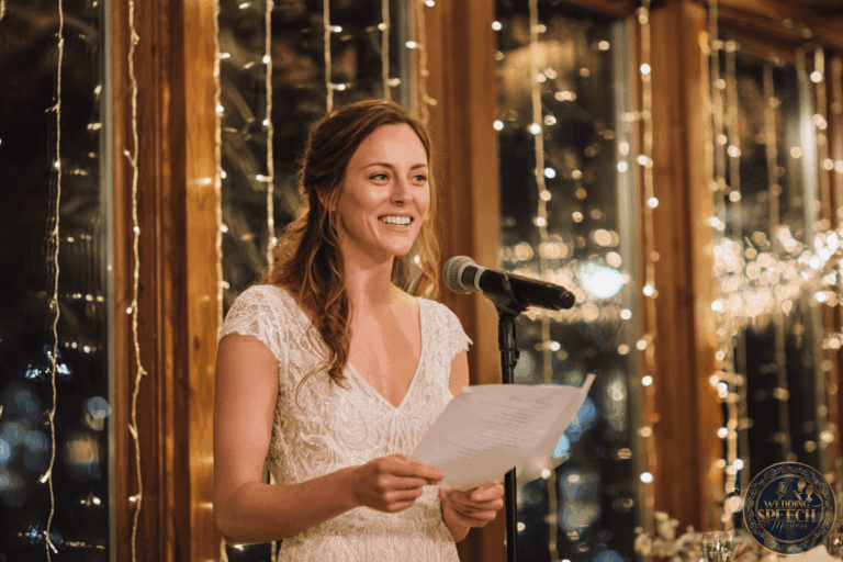 A woman in a white dress stands at a microphone, holding a sheet of paper, speaking in front of large windows decorated with string lights.