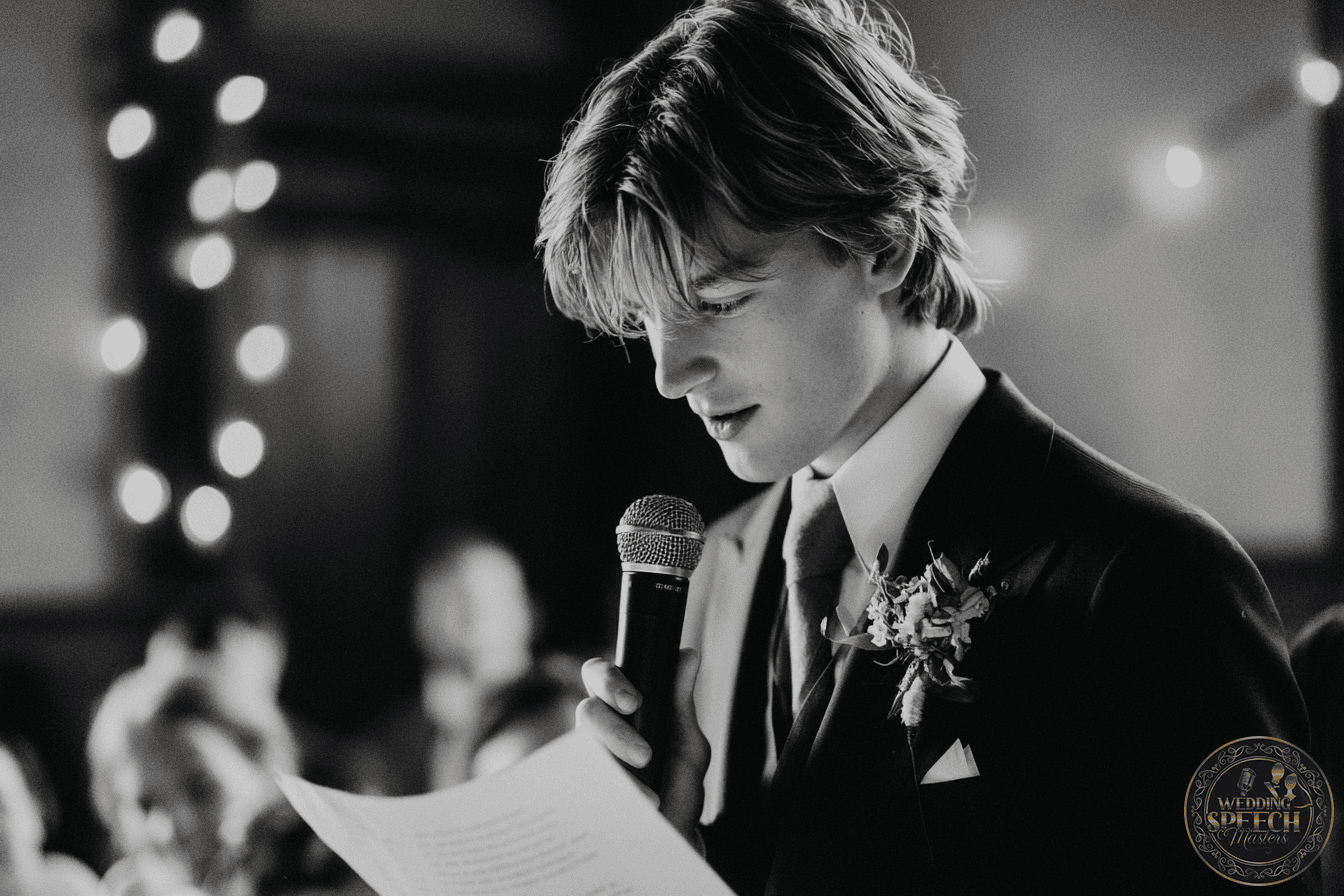 A young man in a suit holds a microphone and reads from a paper, delivering a speech at an indoor event with string lights in the background. For more details or inquiries, please Contact Us.